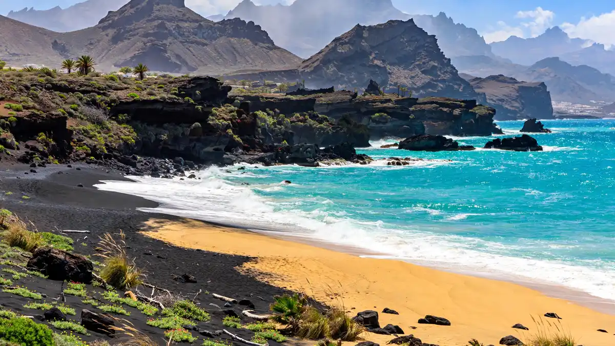 Volcanic Canary Island coastline with a golden and black-sand beach, turquoise Atlantic water and volcanic peaks behind