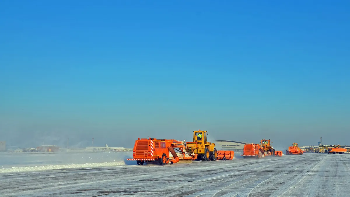 Orange snow-clearing vehicles ploughing a snow-covered airport runway under a clear blue winter sky, with a parked aircraft and terminal buildings in the distance