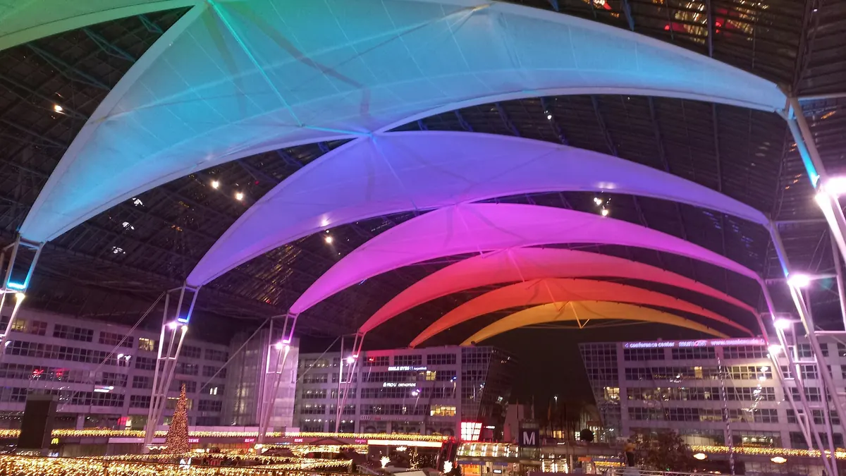 Munich Airport Christmas market at night, with colour-lit canopy sails, festive string lights and a Christmas tree in the forum between the terminals