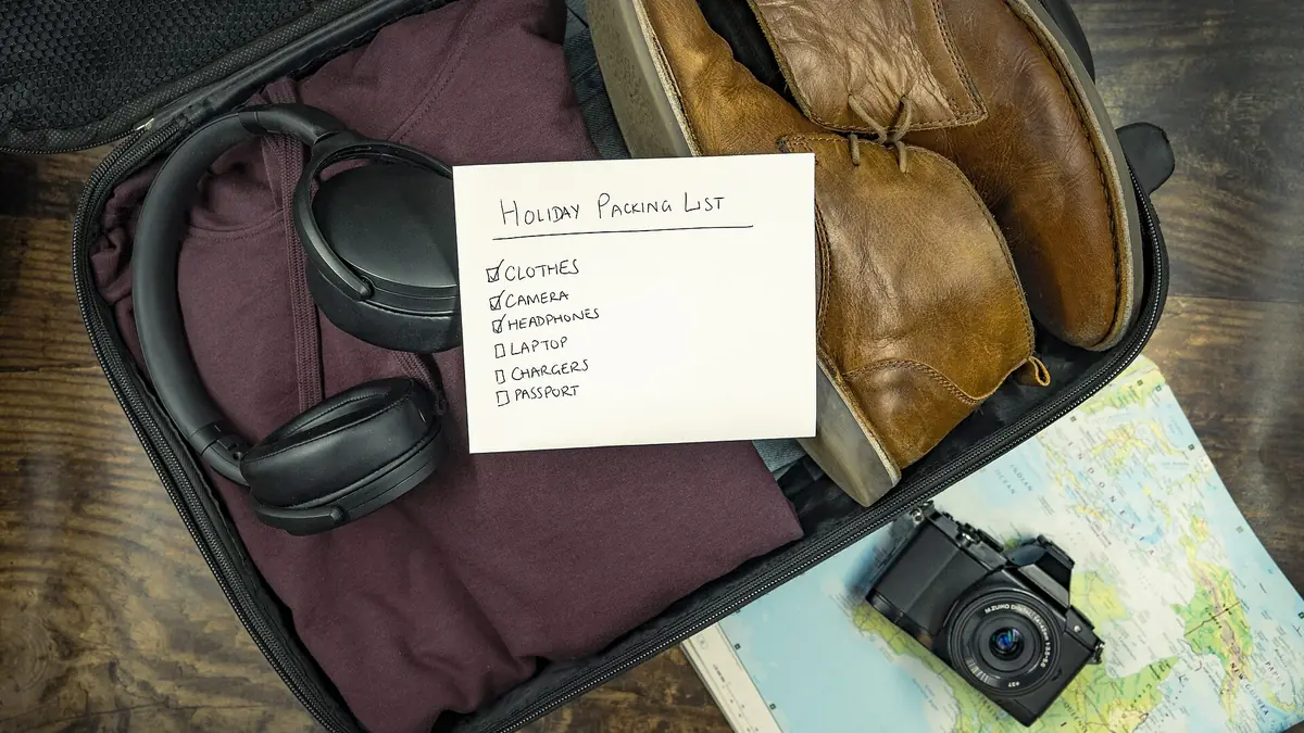 Open suitcase packed for a holiday with folded clothes, headphones, brown shoes and a handwritten packing list, with a camera resting on a map beside it