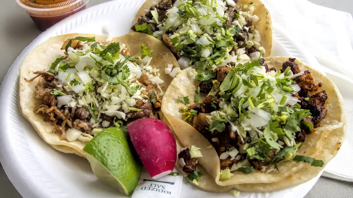 Three tacos on a plate with carnitas, carne asada and al pastor fillings, with lime and salsas