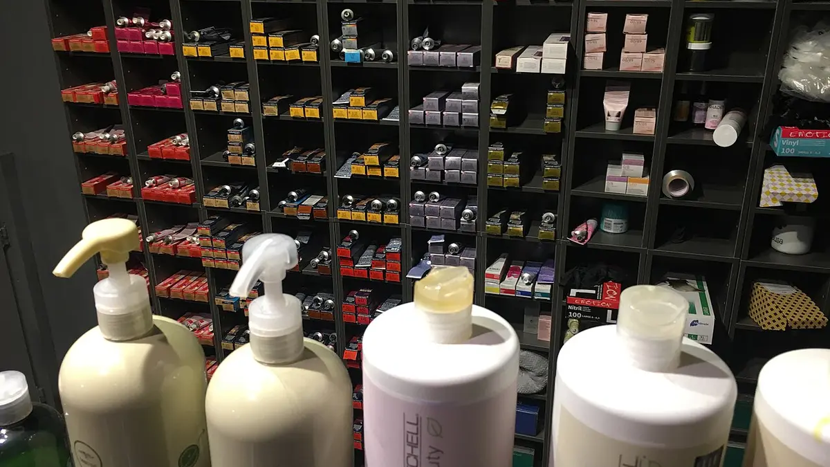 Large hair care bottles in front of tall salon shelves stocked with rows of hair dye boxes, showing the hair care and colour shelves of a beauty supply store