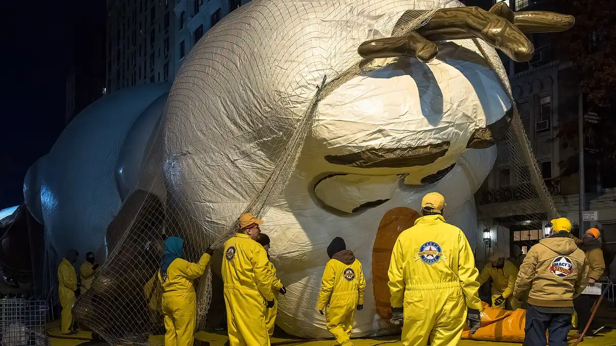 A giant Olaf balloon being inflated at night for the Macy's Thanksgiving Day Parade in New York, held down by netting while crew members in yellow suits work around it