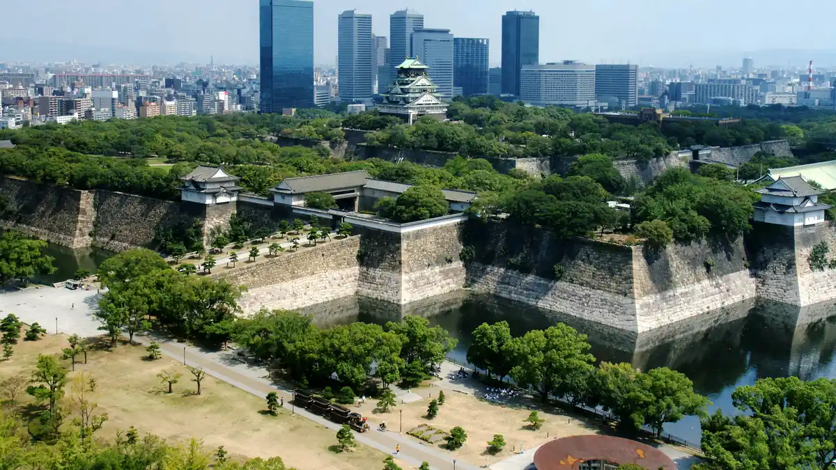 Osaka Castle rising above its stone walls and moat, surrounded by deep green summer trees under a bright sky