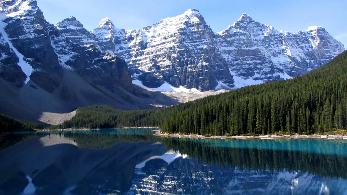 Moraine Lake in Banff National Park with calm turquoise water, pine forest and Rocky Mountain peaks under a clear sky
