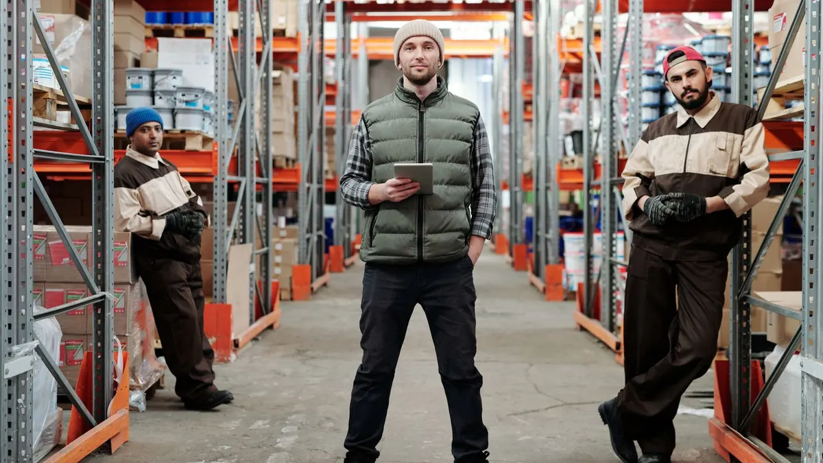 Three warehouse workers standing in a racking aisle, the central worker holding a tablet