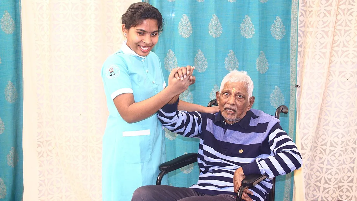 A smiling home carer holding the hand of a senior man seated in a wheelchair at home